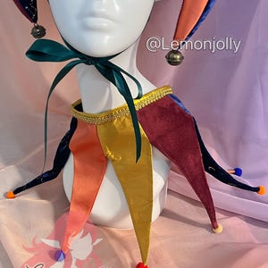 May include: A jester hat and collar set featuring a multi-colored design with velvet and satin fabrics. The hat has dark blue star-patterned points with bells, while the collar features gold, orange, and burgundy points with colorful pom-poms. Text reads @Lemonjolly.