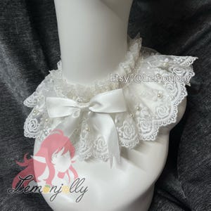 May include: A white lace collar featuring a scalloped edge with floral embroidery, small faux pearls, and a central satin ribbon bow. The collar is displayed on a mannequin neck and includes the text Etsy: @Lemonjolly and a logo.