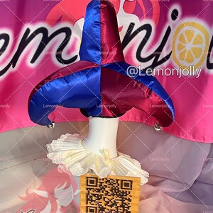 May include: A blue and burgundy jester hat with silver bells, paired with a cream-colored ruffled collar. The hat is displayed on a white mannequin head. A QR code and text are visible in the foreground.