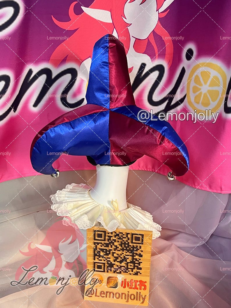 May include: A blue and burgundy jester hat with silver bells, paired with a cream-colored ruffled collar. The hat is displayed on a white mannequin head. A QR code and text are visible in the foreground.