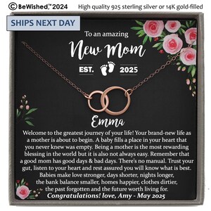 May include: A rose gold-toned necklace featuring two interlocking circular rings, presented in a gray gift box. The card inside displays floral accents, the text "To an amazing New Mom EST. 2025", the name "Emma", and a long congratulatory message.