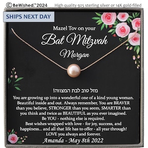 May include: A delicate necklace with a single, round, light pink pearl pendant. The necklace is presented in a gift box with floral accents and text that reads "Mazel Tov on your Bat Mitzvah". The box is black with a silver border.