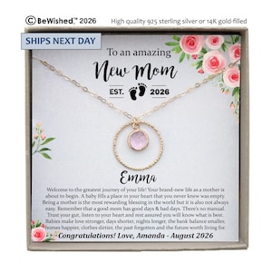 May include: A gold-toned necklace with a circular pendant featuring a small, round, light pink gemstone. The necklace is presented on a card with floral illustrations, the text "To an amazing New Mom EST. 2026", the name "Emma", and a printed message.