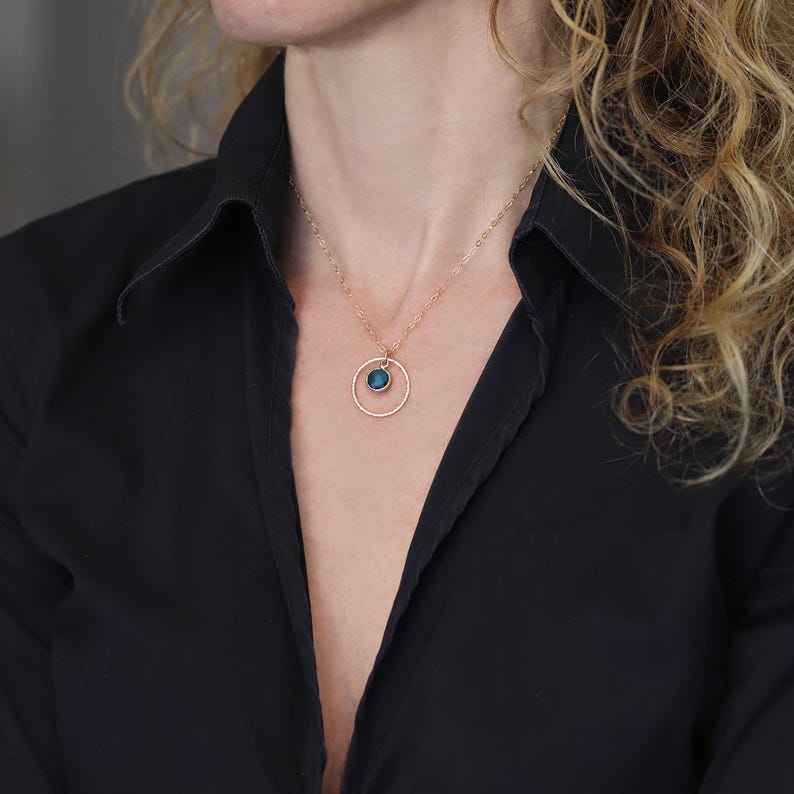 Puede incluir: Un collar de tono dorado con una cadena delicada y un colgante circular. El colgante consiste en un anillo fino y texturizado de tono dorado que enmarca una peque&ntilde;a piedra preciosa redonda de color azul profundo engastada en el centro superior.