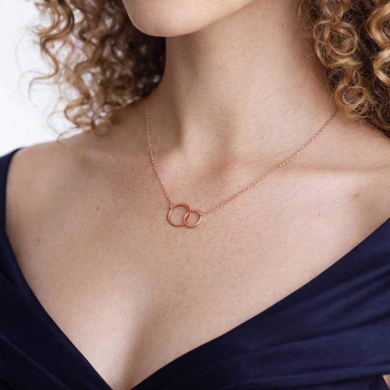 May include: A rose gold necklace featuring two interlocking circular rings as a central pendant. The delicate chain is secured around the neck, with the two rings resting against the chest. The metal has a polished finish and a warm pinkish hue.