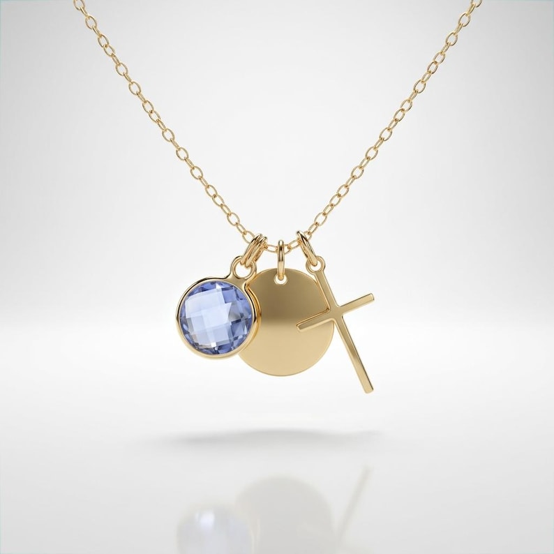 May include: A gold chain necklace featuring three dangling charms: a round faceted light blue gemstone, a plain circular gold disc, and a simple gold cross. The necklace is constructed with a delicate link chain and polished gold metal components.