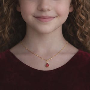 Puede incluir: Un collar dorado con un peque&ntilde;o colgante rojo en forma de l&aacute;grima. El collar lo lleva una persona con una prenda de vestir de color rojo oscuro. La cadena del collar tiene peque&ntilde;as cuentas espaciadas.