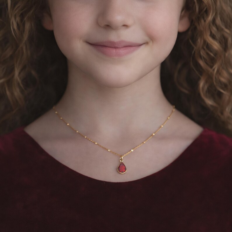 Puede incluir: Un collar dorado con un peque&ntilde;o colgante rojo en forma de l&aacute;grima. El collar lo lleva una persona con una prenda de vestir de color rojo oscuro. La cadena del collar tiene peque&ntilde;as cuentas espaciadas.