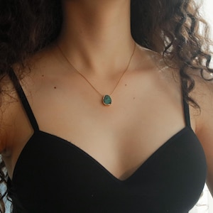 May include: A gold-toned necklace with a teal-colored pendant. The pendant has a unique, irregular shape and is set in a gold bezel. The necklace is worn against a black top, creating a contrast that highlights the jewelry.