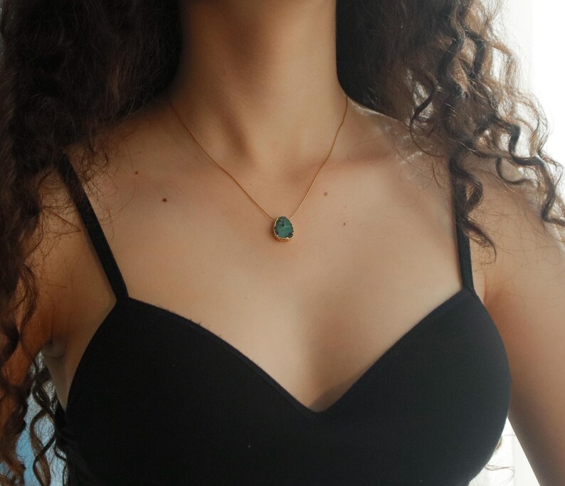 May include: A gold-toned necklace with a teal-colored pendant. The pendant has a unique, irregular shape and is set in a gold bezel. The necklace is worn against a black top, creating a contrast that highlights the jewelry.