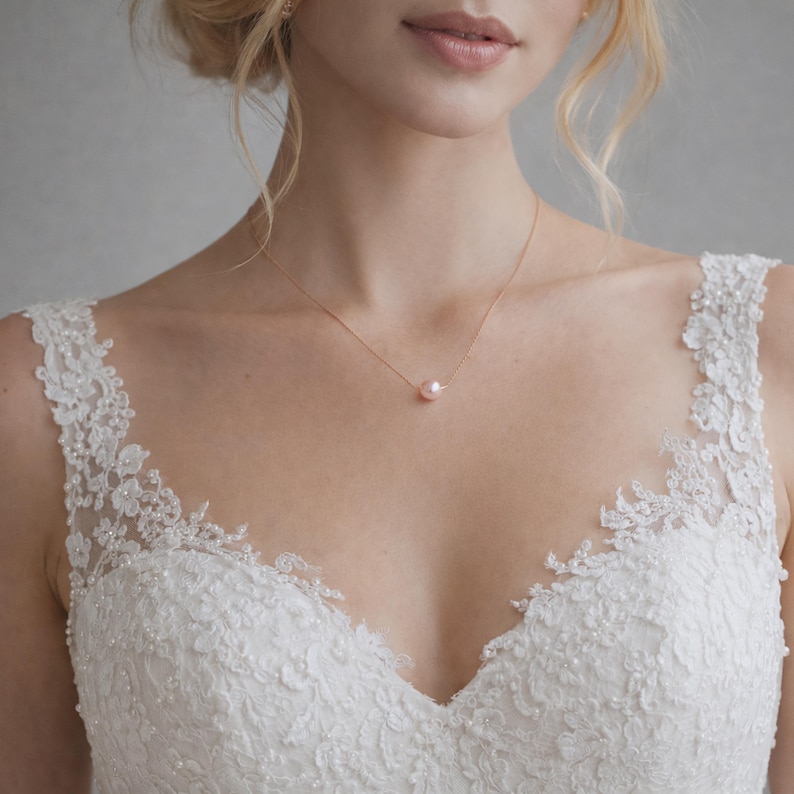 May include: A delicate rose gold necklace featuring a single, soft pink pearl pendant. The necklace is displayed on a person wearing a white lace wedding dress, creating an elegant and romantic aesthetic. The pearl is approximately 0.3 inches in diameter.