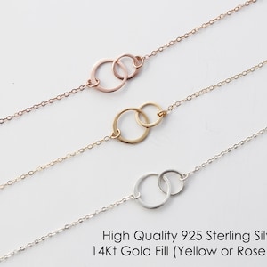Puede incluir: Tres collares de cadena delicada, cada uno con dos anillos circulares entrelazados como colgante central. El collar superior es de oro rosa, el del medio es de oro amarillo y el inferior es de plata. Texto: High Quality 925 Sterling Silver or 14Kt Gold Fill (Yellow or Rose Gold).