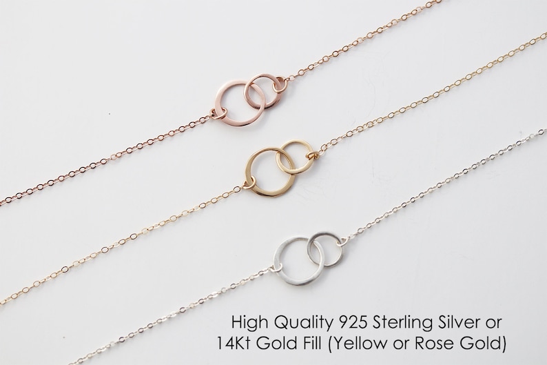 Puede incluir: Tres collares de cadena delicada, cada uno con dos anillos circulares entrelazados como colgante central. El collar superior es de oro rosa, el del medio es de oro amarillo y el inferior es de plata. Texto: High Quality 925 Sterling Silver or 14Kt Gold Fill (Yellow or Rose Gold).