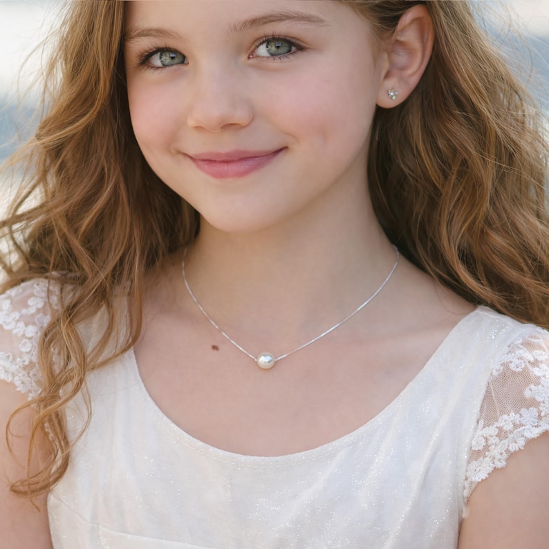 Puede incluir: Un collar de plata con un colgante de perla blanca, redondo y grande. El collar se lleva sobre un vestido blanco con detalles de encaje. La imagen es un retrato de una persona con cabello largo, ondulado, casta&ntilde;o claro y ojos azules.