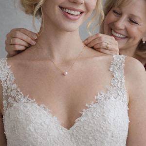 May include: A gold necklace with a single pearl pendant is being clasped around a person's neck. The person is wearing a white lace dress. Another person is smiling and helping to fasten the necklace.