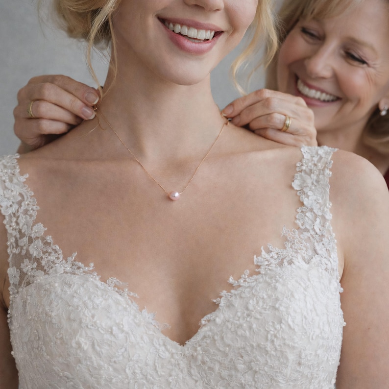May include: A gold necklace with a single pearl pendant is being clasped around a person's neck. The person is wearing a white lace dress. Another person is smiling and helping to fasten the necklace.