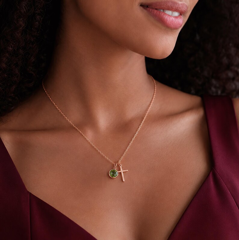 May include: A rose gold chain necklace featuring two small charms: a round, bezel-set green gemstone and a simple, plain cross pendant. The delicate metal chain rests against the neck, showcasing the two distinct charms hanging together.