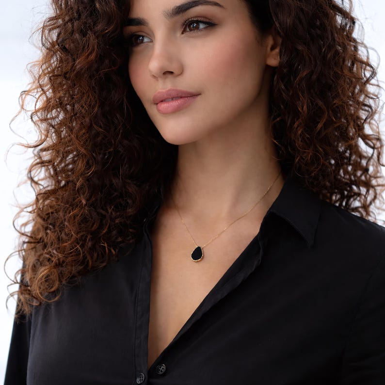 May include: A gold necklace with a black teardrop-shaped pendant. The pendant is set in a gold frame and hangs from a delicate gold chain. The necklace is worn against a black collared shirt.