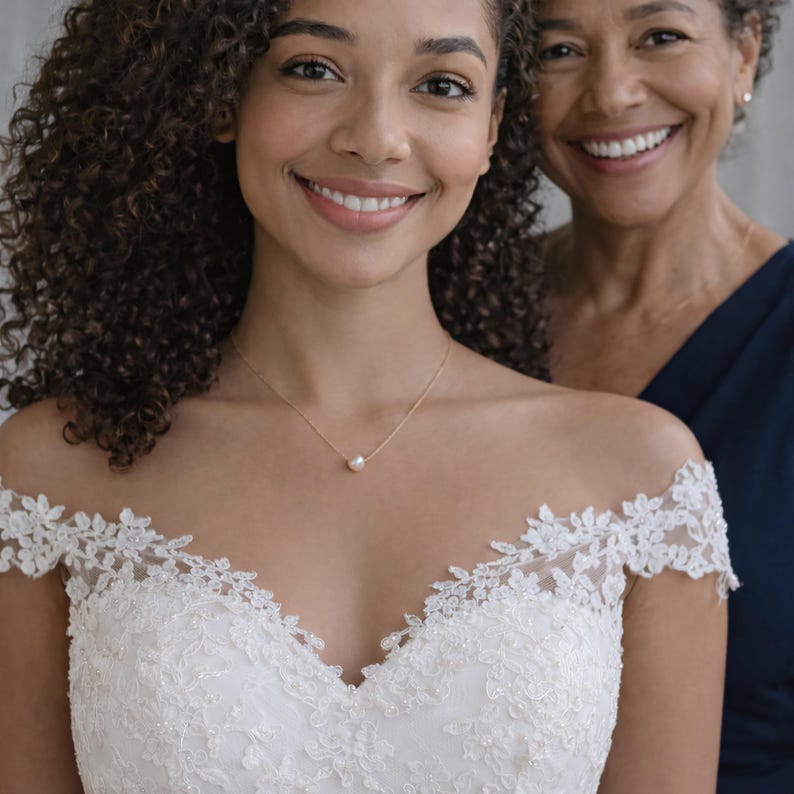 May include: A woman wearing a white off-the-shoulder wedding dress with lace detailing. She is wearing a delicate gold necklace with a single pearl pendant. Another woman in a navy blue dress is smiling in the background.
