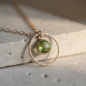 Puede incluir: Un collar de oro con un colgante circular de superficie texturizada. Una piedra preciosa de peridoto redonda, facetada y de color verde claro est&aacute; engastada en el centro del c&iacute;rculo de oro. La cadena est&aacute; formada por finos eslabones dorados.