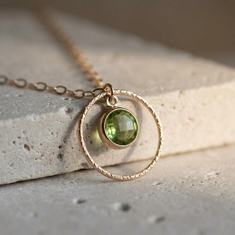 Puede incluir: Un collar de oro con un colgante circular de superficie texturizada. Una piedra preciosa de peridoto redonda, facetada y de color verde claro est&aacute; engastada en el centro del c&iacute;rculo de oro. La cadena est&aacute; formada por finos eslabones dorados.