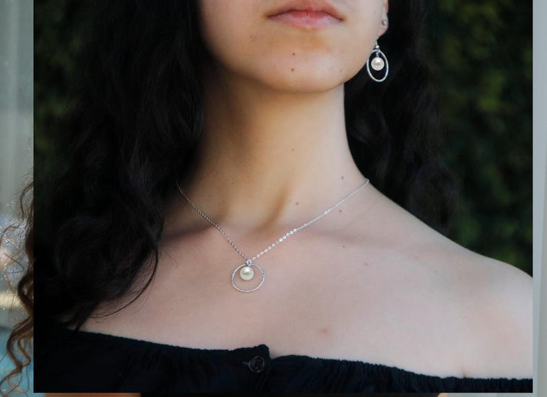 May include: A silver necklace with a pearl pendant and a circle design. The necklace is worn by a person with long dark hair.