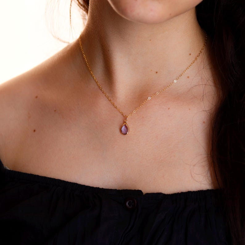 Puede incluir: Un collar dorado con un peque&ntilde;o colgante de piedra preciosa p&uacute;rpura en forma de l&aacute;grima. El collar es delicado y parece ser usado por una persona. El fondo es de un color suave y neutro, resaltando la joya.