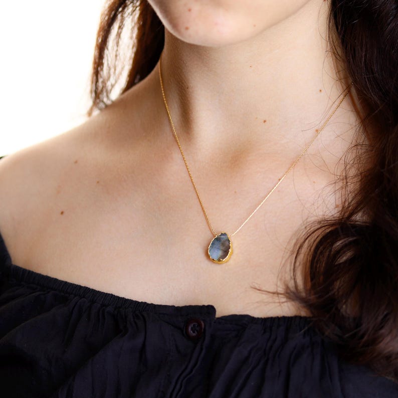 May include: A gold-colored necklace with a teardrop-shaped pendant. The pendant has a light blue and gray stone with a gold-colored border. The necklace is worn on a person with dark hair and a black top.