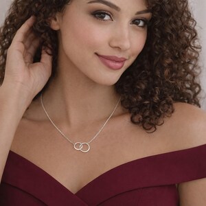 May include: A silver necklace featuring two small, interlocking circular pendants suspended from a delicate chain. The necklace rests against the collarbone, showcasing a polished metallic finish and a simple, clean design with two connected rings.
