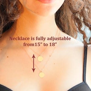 May include: A gold-toned necklace featuring two delicate chains, each with a small, circular pendant. The text on the image states: Necklace is fully adjustable from 15" to 18". A vertical arrow indicates the adjustable length of the chain.