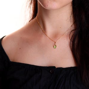 Puede incluir: Un collar dorado con un peque&ntilde;o colgante de piedra preciosa verde claro en forma de pera. La cadena del collar es delicada y tiene peque&ntilde;as cuentas espaciadas uniformemente. El colgante es de un color verde vibrante. El collar se lleva sobre una blusa negra.