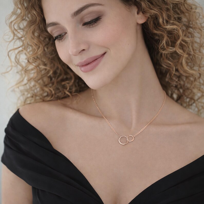 May include: A rose gold necklace featuring two interlocking circular rings as a pendant on a delicate chain. The metal has a polished finish, and the design consists of one larger ring connected to a smaller ring, suspended from a thin link chain.