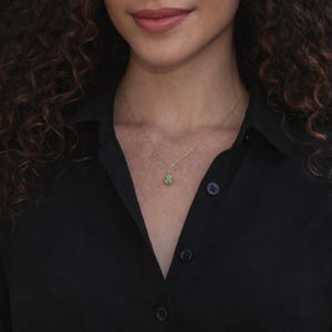 Puede incluir: Un collar de oro con un peque&ntilde;o colgante de piedra verde en forma de l&aacute;grima. El collar es delicado y cuelga de una cadena de oro. La persona lleva una camisa negra abotonada.