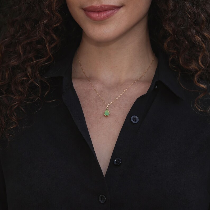 Puede incluir: Un collar de oro con un peque&ntilde;o colgante de piedra verde en forma de l&aacute;grima. El collar es delicado y cuelga de una cadena de oro. La persona lleva una camisa negra abotonada.