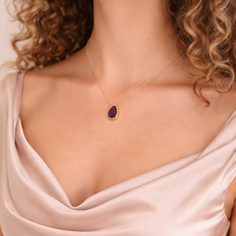 May include: A gold-toned necklace featuring a teardrop-shaped, deep purple gemstone pendant. The pendant is set in a gold bezel. The necklace is worn against a backdrop of a light pink satin fabric.
