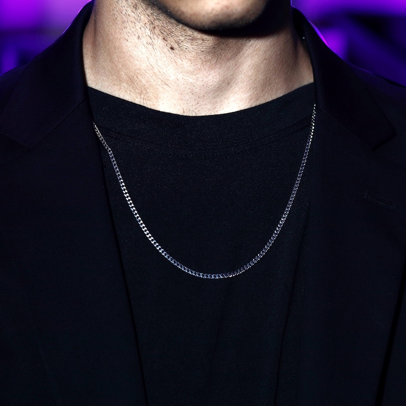 May include: A thin, silver-toned metal chain necklace with small, interlocking links. The necklace is worn around the neck, resting against a plain black t-shirt and a black blazer, featuring a simple, uniform design throughout the entire length.