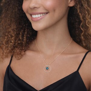 May include: A gold-toned necklace featuring a delicate chain and a circular pendant. The pendant consists of a thin, textured gold-toned ring framing a small, round, deep teal-colored gemstone set in the center. The necklace rests against the neck.
