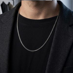 May include: A thin, silver-colored metal chain necklace with small, interlocking links. The necklace rests against a plain black t-shirt, which is worn underneath a dark gray, textured blazer. The chain has a polished, reflective finish.