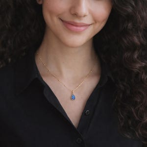 Puede incluir: Un collar dorado con un peque&ntilde;o colgante en forma de l&aacute;grima. El colgante tiene un centro azul oscuro y un marco dorado. El collar se lleva sobre una camisa negra.
