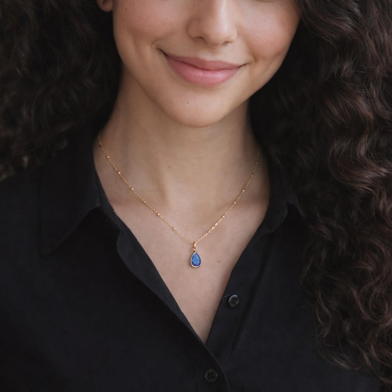 Puede incluir: Un collar dorado con un peque&ntilde;o colgante en forma de l&aacute;grima. El colgante tiene un centro azul oscuro y un marco dorado. El collar se lleva sobre una camisa negra.