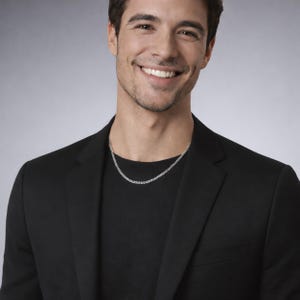 May include: A silver curb chain necklace featuring interlocking flat links with a polished finish. The necklace rests against a black t-shirt worn underneath a black blazer, showcasing a classic metal design that sits close to the base of the neck.