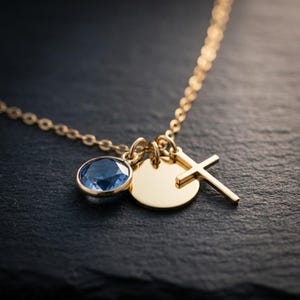 May include: A gold-toned necklace featuring three charms: a round, faceted blue gemstone in a bezel setting, a plain circular gold-toned disc, and a simple gold-toned cross pendant, all suspended from a delicate gold-toned link chain.