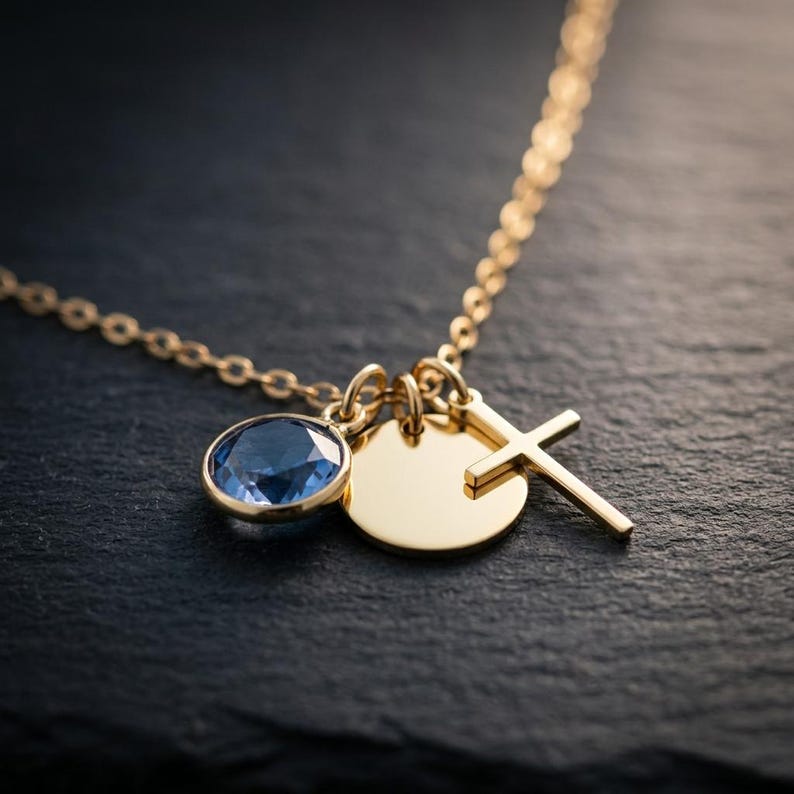 May include: A gold-toned necklace featuring three charms: a round, faceted blue gemstone in a bezel setting, a plain circular gold-toned disc, and a simple gold-toned cross pendant, all suspended from a delicate gold-toned link chain.