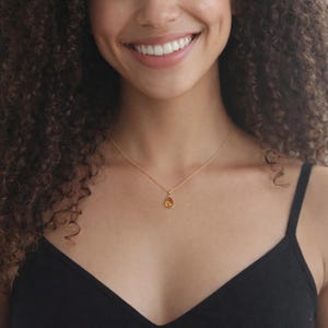 Puede incluir: Un collar dorado con un peque&ntilde;o colgante en forma de l&aacute;grima. El colgante es de color &aacute;mbar c&aacute;lido y cuelga de una delicada cadena dorada. El collar se lleva sobre una camiseta negra.
