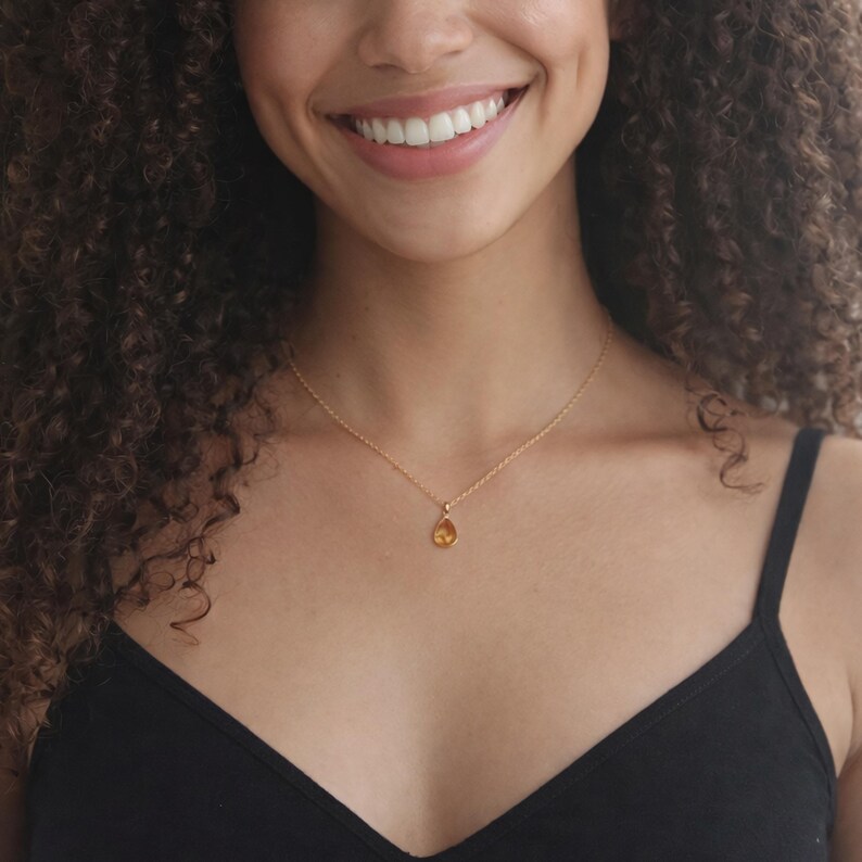 Puede incluir: Un collar dorado con un peque&ntilde;o colgante en forma de l&aacute;grima. El colgante es de color &aacute;mbar c&aacute;lido y cuelga de una delicada cadena dorada. El collar se lleva sobre una camiseta negra.