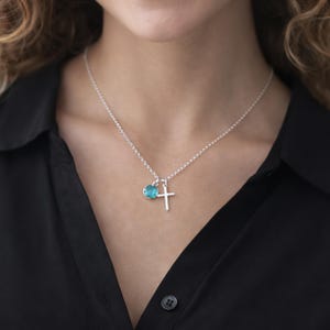 Puede incluir: Un collar de plata con un peque&ntilde;o colgante de cruz y un charm redondo de piedra preciosa azul claro. El collar se lleva sobre una camisa negra.