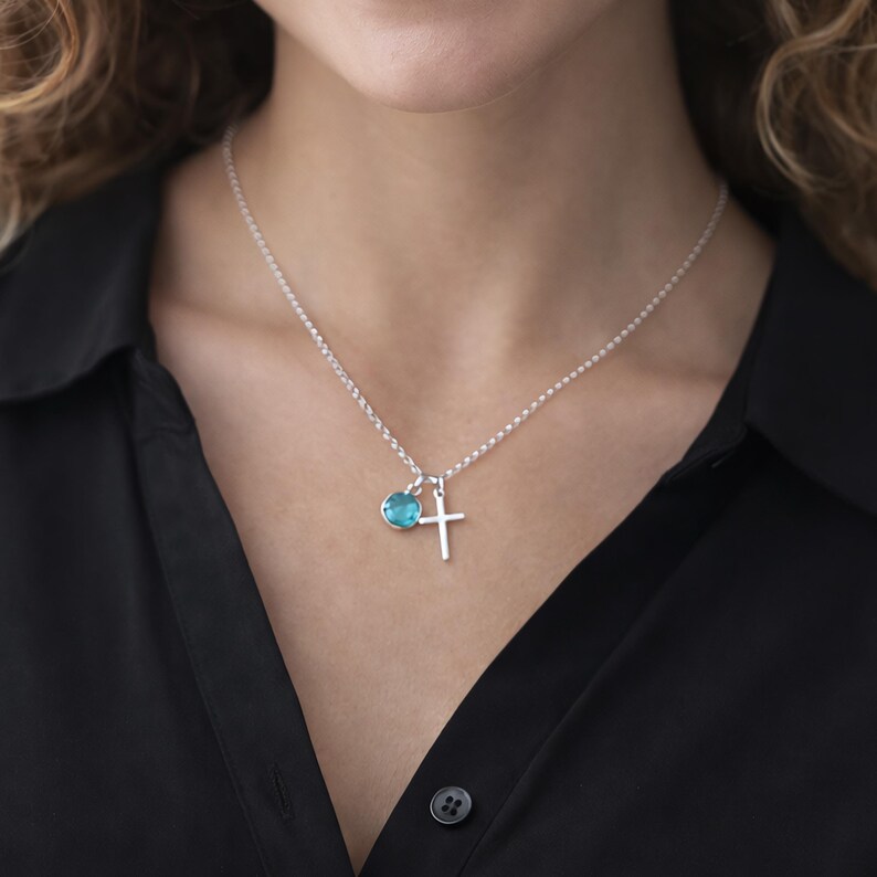 Puede incluir: Un collar de plata con un peque&ntilde;o colgante de cruz y un charm redondo de piedra preciosa azul claro. El collar se lleva sobre una camisa negra.