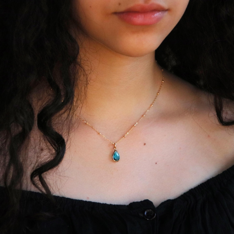 Puede incluir: Un collar dorado con una cadena delicada y un colgante en forma de l&aacute;grima. El colgante es de color turquesa vibrante, engastado en un bisel dorado. El collar se est&aacute; usando.