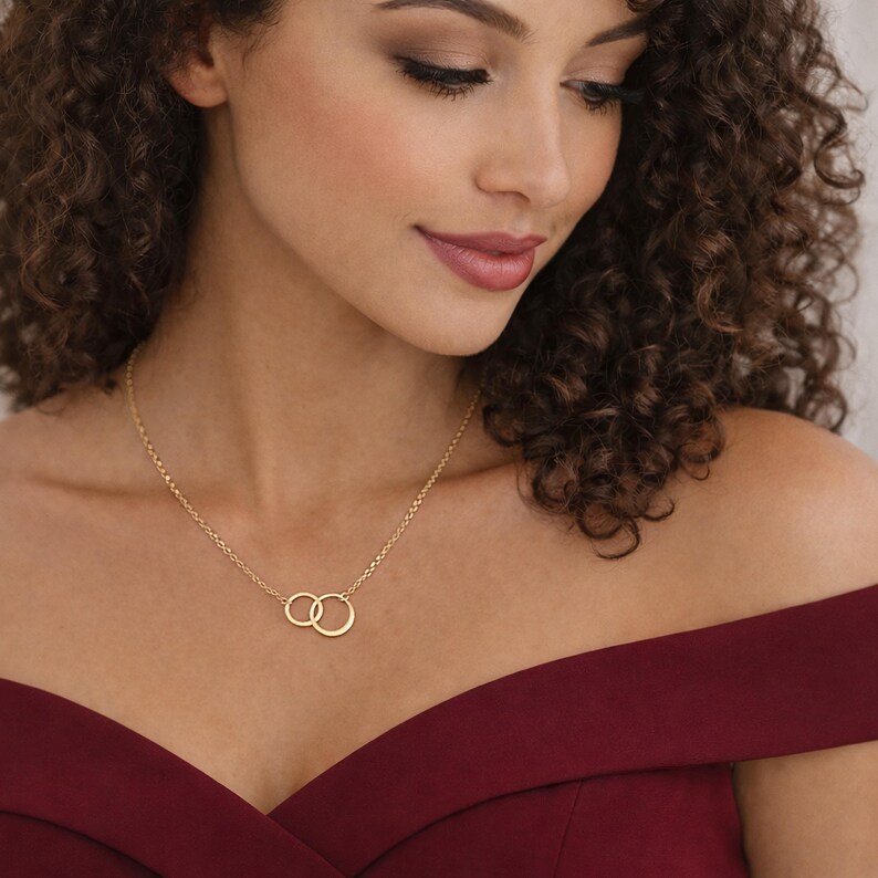 May include: A gold-toned necklace featuring two small, interlocking circular rings as a central pendant. The delicate chain is secured around the neck, with the two rings resting against the collarbone area, creating a simple and clean design.