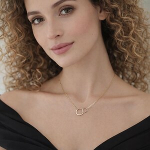 May include: A gold-toned necklace featuring two interlocking circular rings as a central pendant. The pendant is suspended from a delicate, thin gold-toned chain that rests against the collarbone. The design consists of two smooth, polished rings.